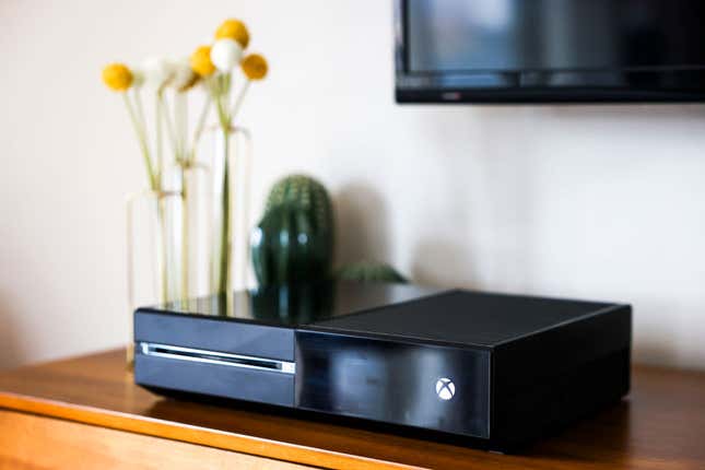 An Xbox One console sits on a wooden cabinet with a vase and flowers in the background.