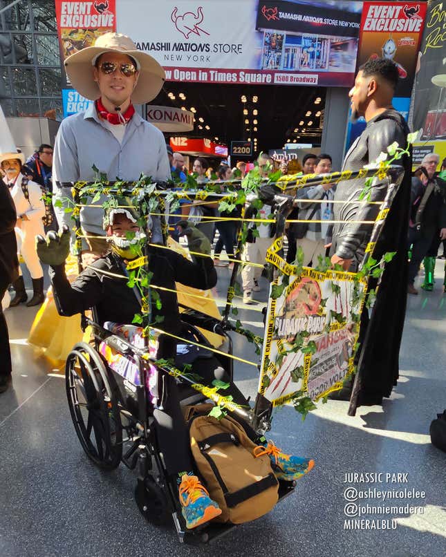 A cosplayer dressed as Jurassic Park's Alan Grant wheels a cosplayer in a wheelchair dressed as a velociraptor.